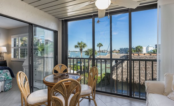 Interior view of a coastal apartment balcony with rattan chairs and table, looking out to a sunny beachscape through large sliding glass doors.