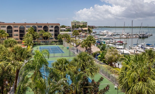 Aerial view of a coastal resort with tennis courts, a marina filled with boats, and surrounding buildings under a clear sky.