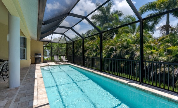 A screened patio featuring a long narrow swimming pool, surrounded by lush greenery, attached to a modern house with a visible outdoor dining area.