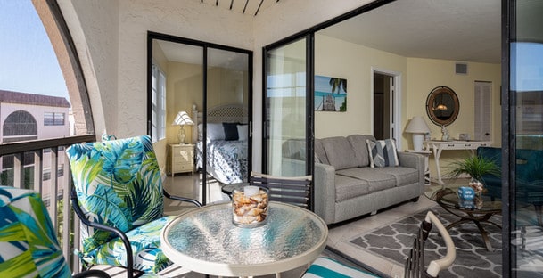 Bright balcony with a round table and snacks, tropical print chairs, overlooking an open-plan living room with modern decor.