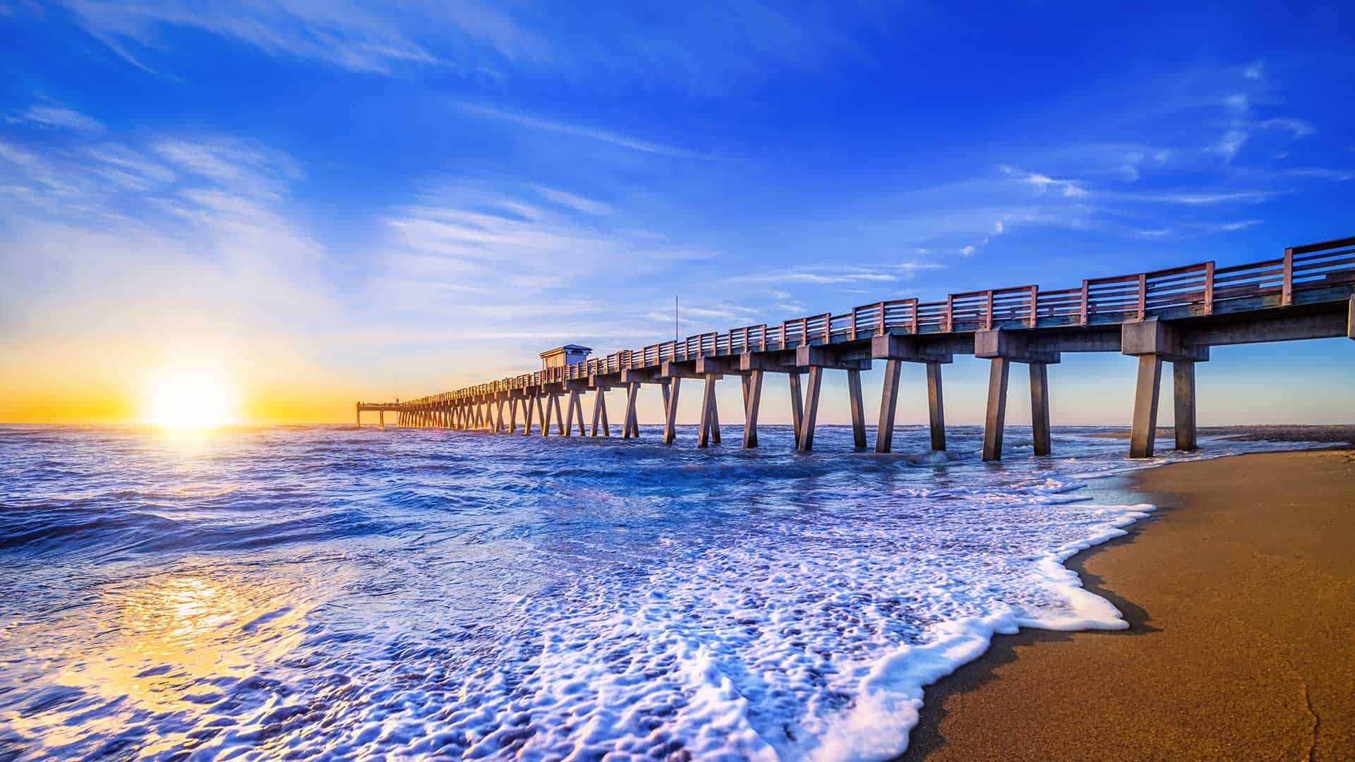 A long pier extends into the sea against a vibrant sunset with light reflecting off gentle waves.